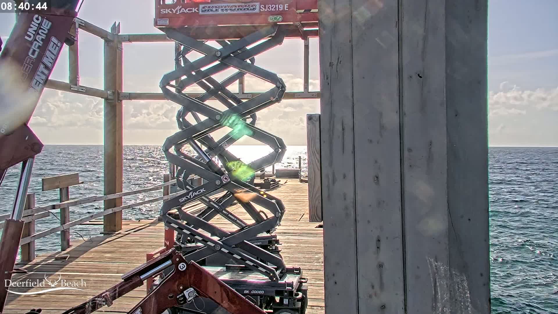 On a sunny day, a partially constructed structure on a wooden pier overlooks a calm ocean, with two scissor lifts positioned near the structure.