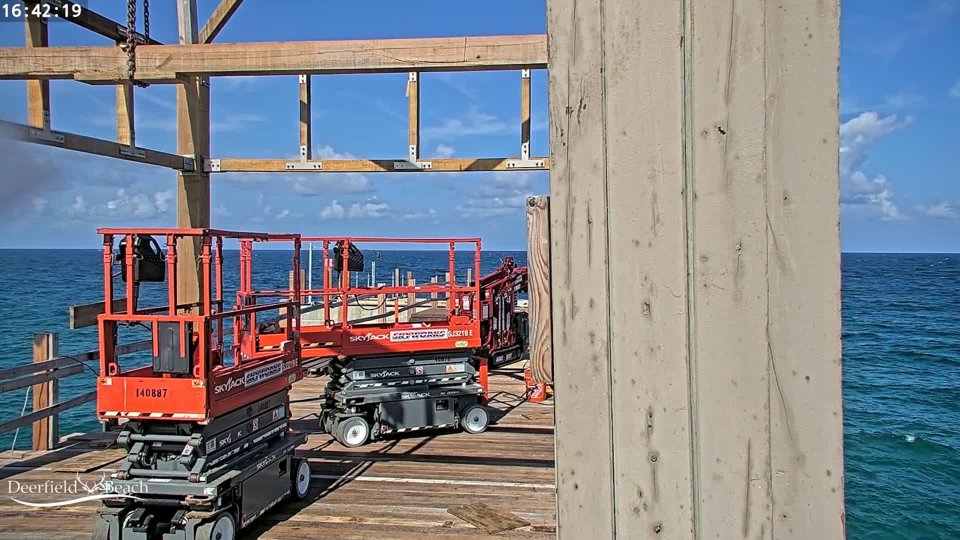 Two red scissor lifts are parked on a wooden pier overlooking a calm ocean under a partly cloudy blue sky.