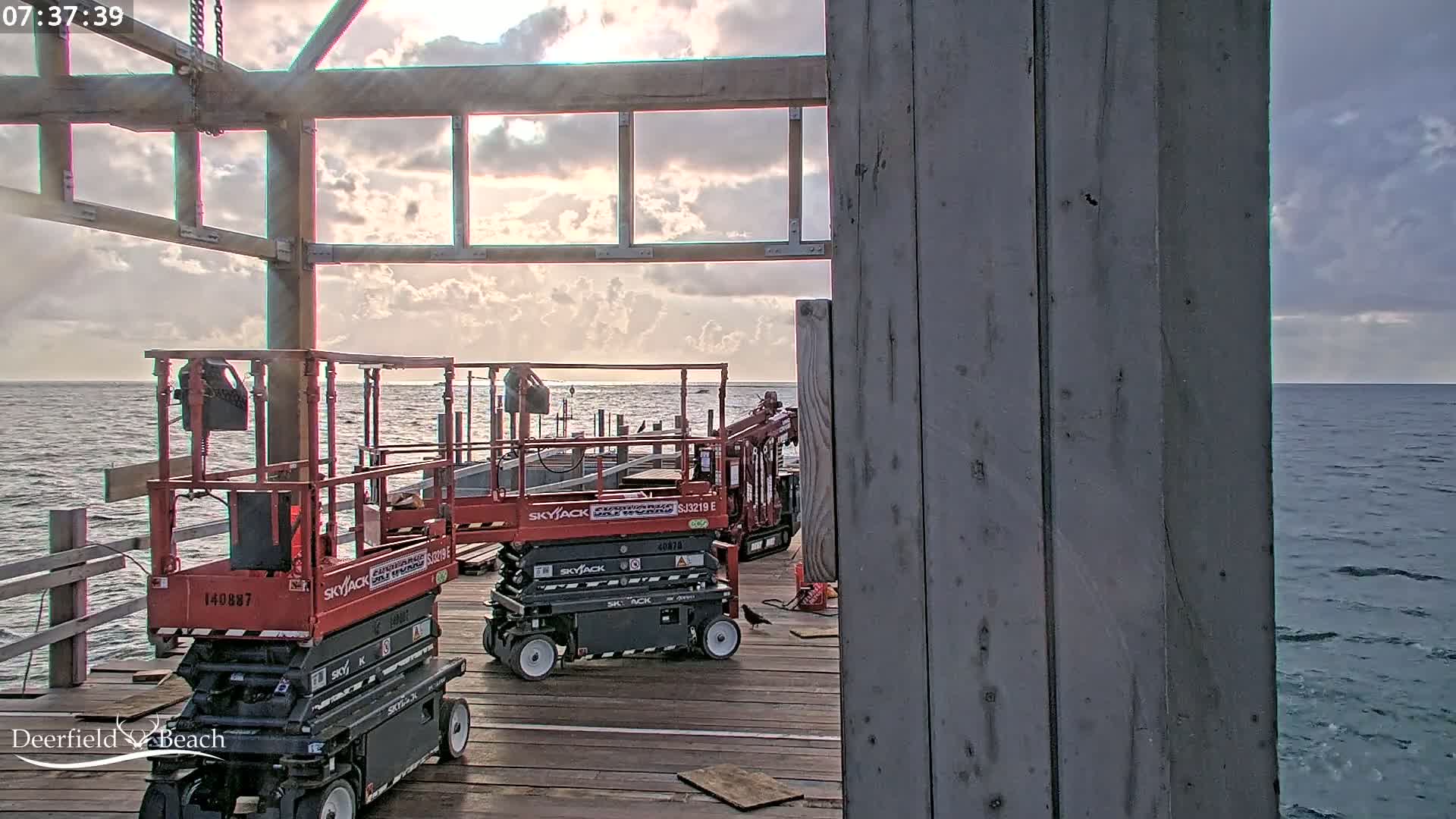 Two red scissor lifts are parked on a wooden pier overlooking a calm ocean under a partly cloudy sky.