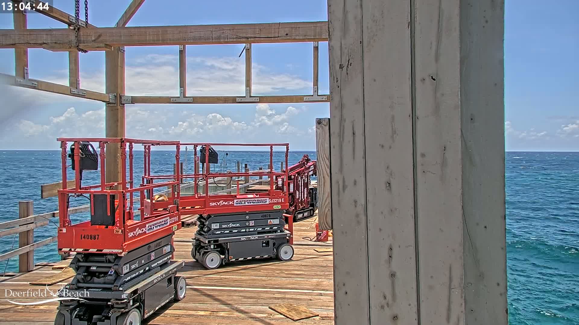 Two red scissor lifts are parked on a wooden deck overlooking a blue ocean under a partly cloudy sky.