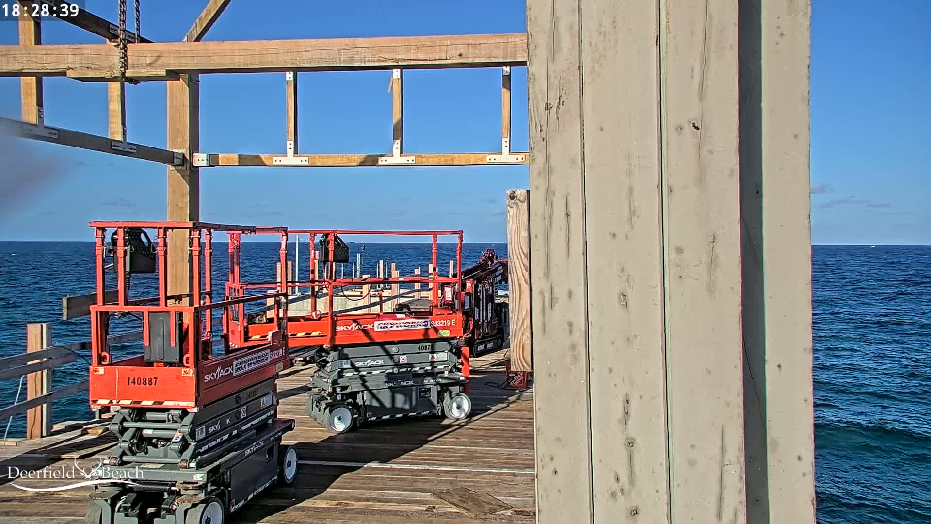 Two red scissor lifts are parked on a wooden deck overlooking a blue ocean under a partly cloudy sky.