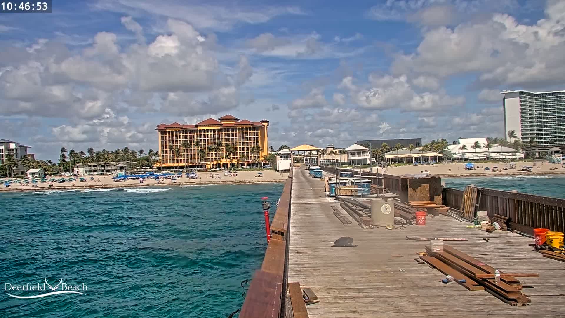 A partly cloudy day at a beach with a long wooden pier extending from the foreground to a sandy shore lined with buildings, including a large yellow hotel.