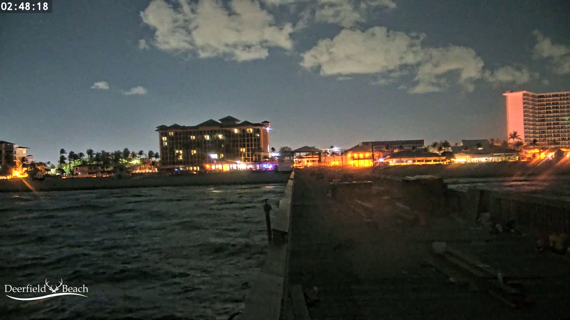 A nighttime view from a pier shows a partly cloudy sky over a dark, choppy ocean with illuminated beachfront buildings.