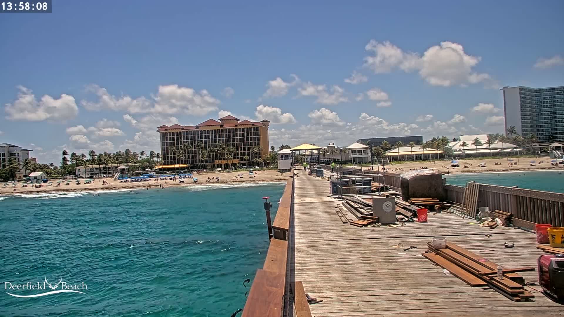 A partly cloudy day shows a wooden pier extending over teal ocean water towards a sandy beach with people and buildings under a mostly sunny sky.