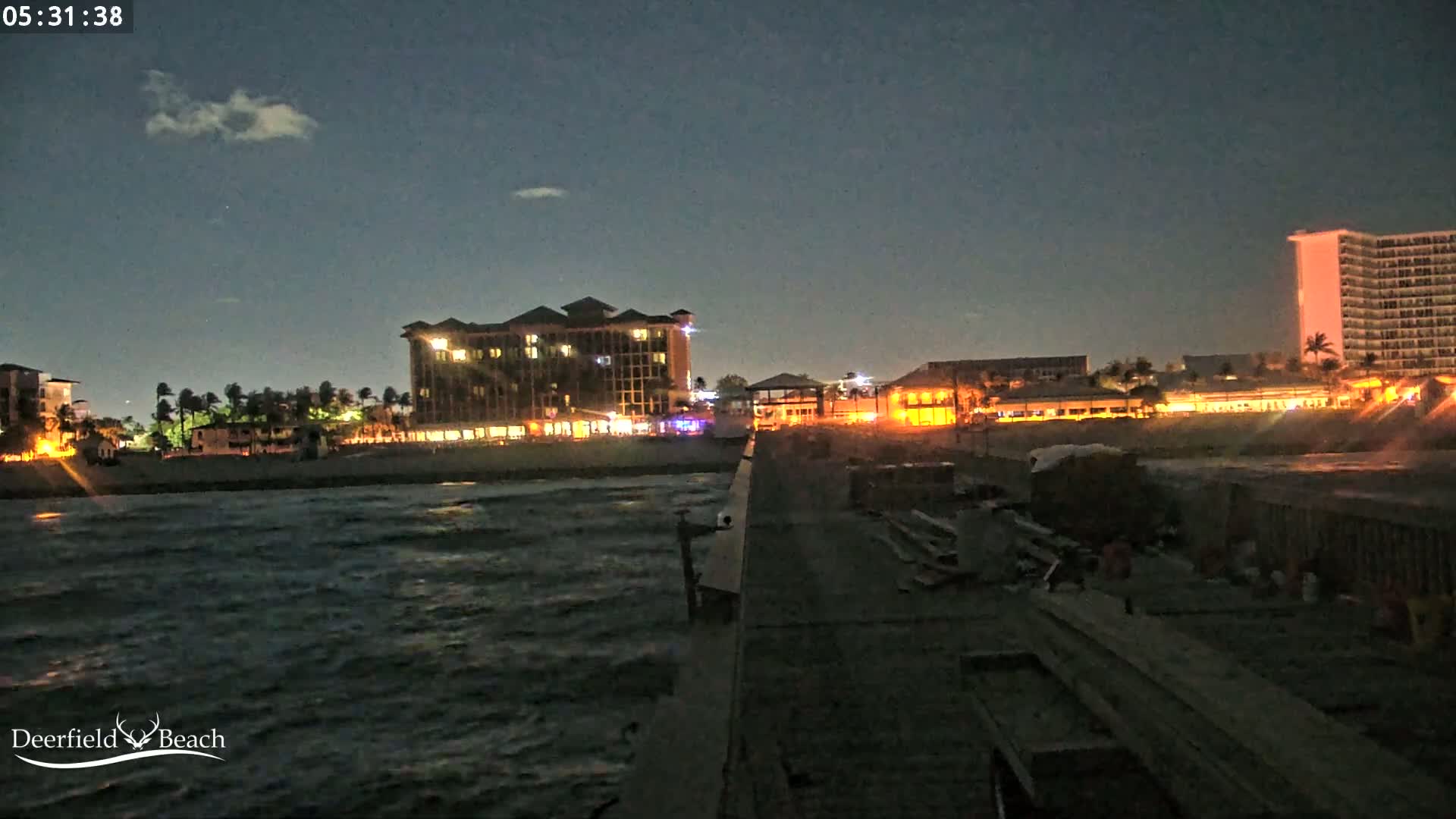 A nighttime view from a pier shows a shoreline with illuminated hotels and buildings, and a slightly choppy ocean.