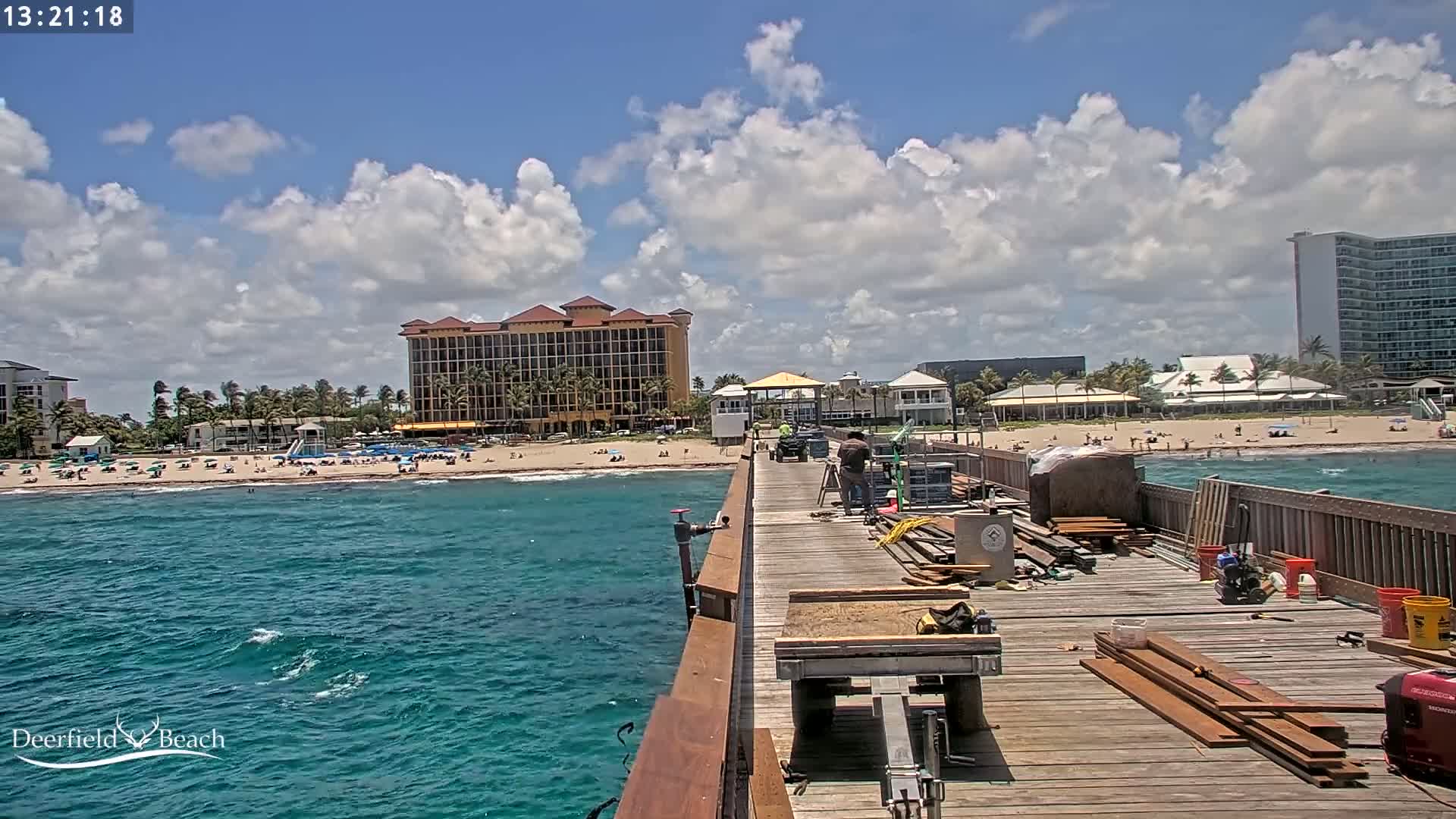 A partly cloudy day shows a wooden pier under construction overlooking a beach with people, hotels, and ocean waves.