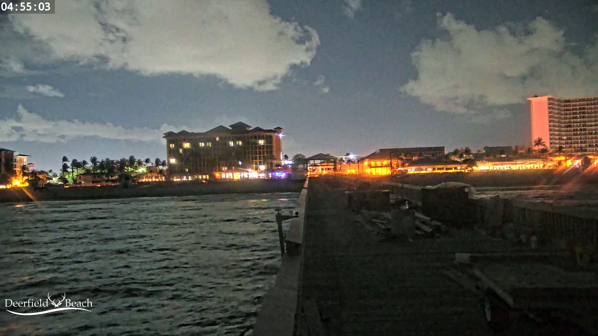 A nighttime view from a pier shows a dark, choppy ocean, illuminated buildings along a beach, and a partly cloudy sky.