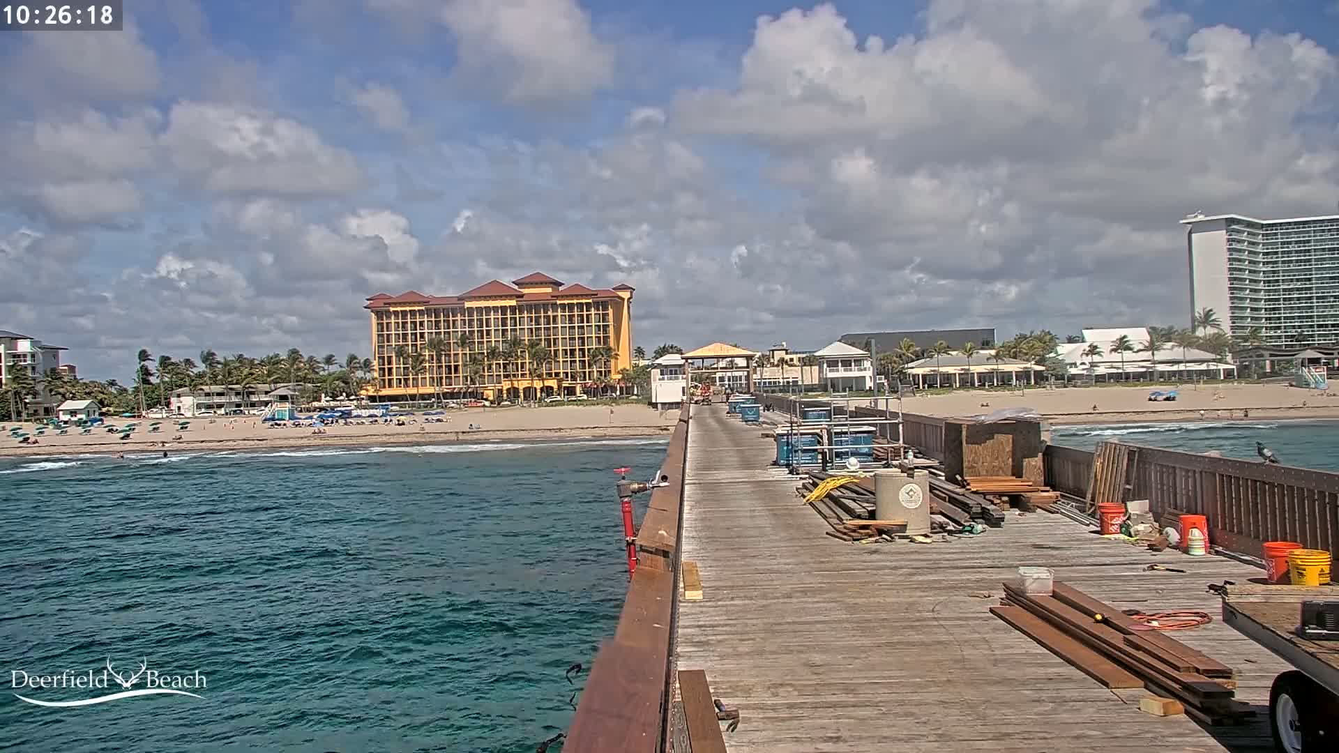 A partly cloudy day shows a wooden pier extending from the shore toward the ocean, with construction materials on the pier and beachgoers visible on the sand in front of beachfront hotels.
