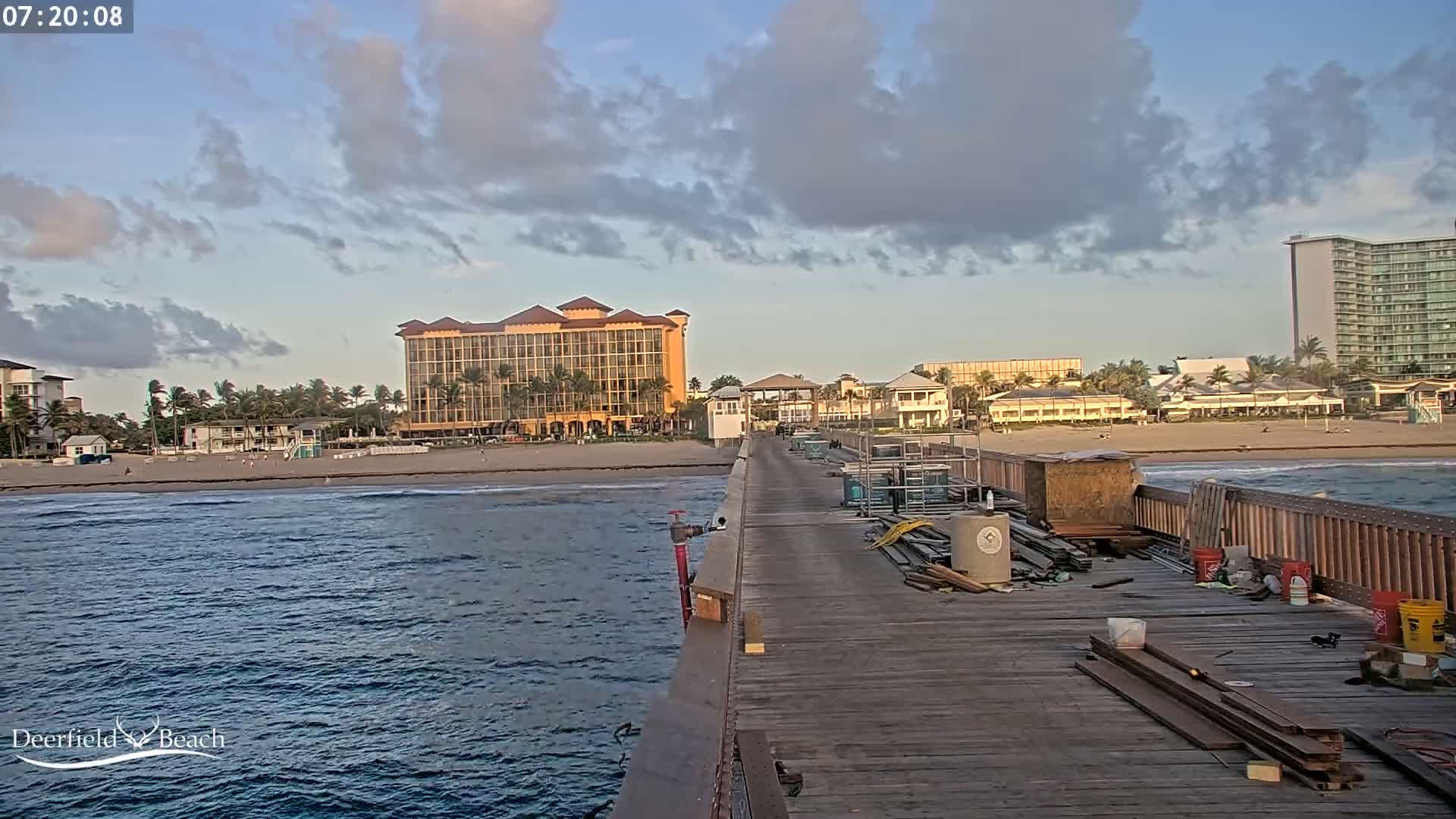 A view from a pier shows a partly cloudy sky over a beach with hotels and buildings, and construction materials are scattered on the pier.