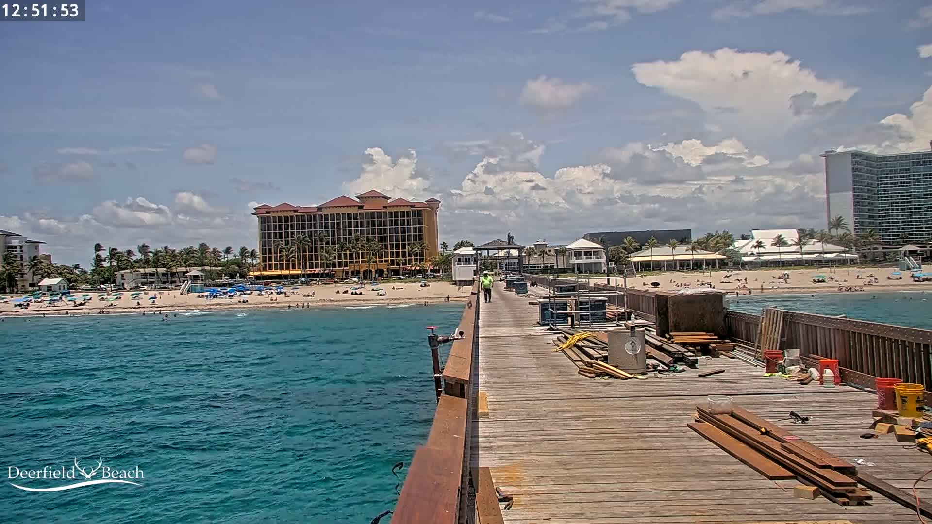 A partly cloudy day shows a wooden pier under construction extending over blue ocean water toward a beach with people and buildings.