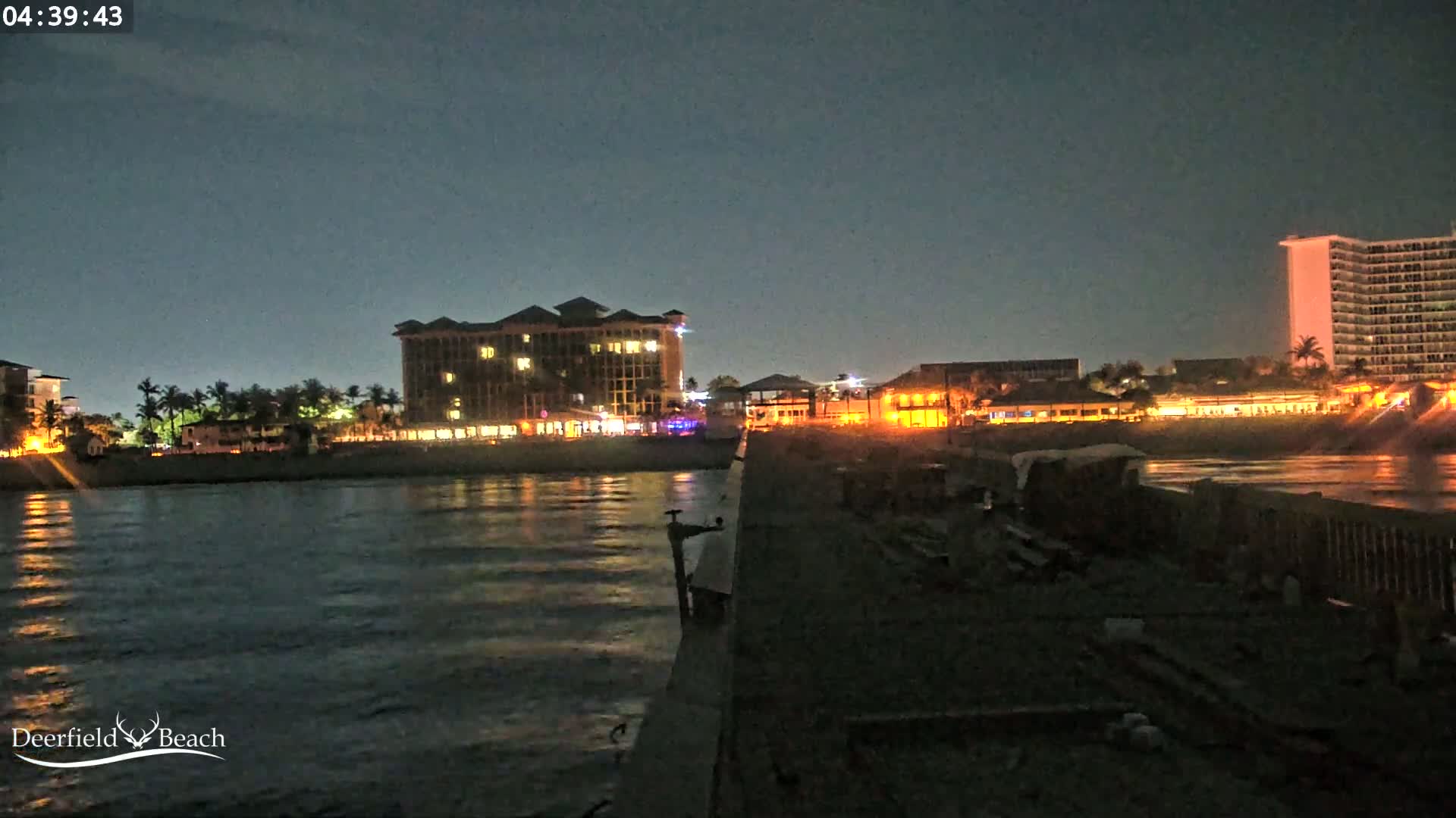 A nighttime view from a pier shows a calm body of water reflecting the lights of buildings along the shore.