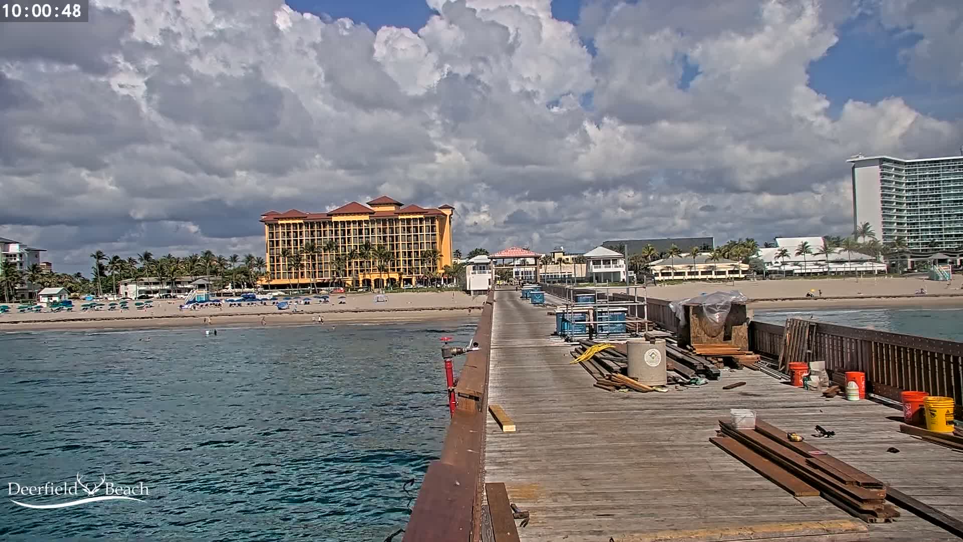 A partly cloudy day shows a view from a pier under construction overlooking a beach with people and several buildings, including a large yellow hotel.