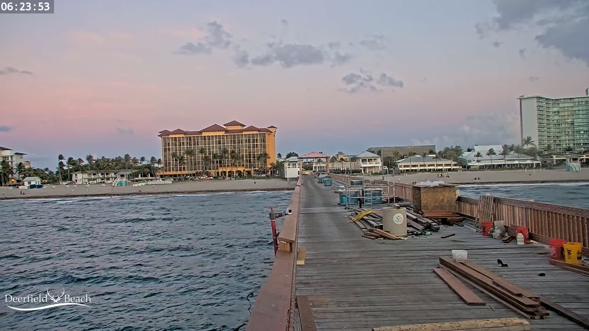 A pier under construction extends into a wavy ocean, with hotels and a beach visible in the background under a partly cloudy, pinkish-purple twilight sky.