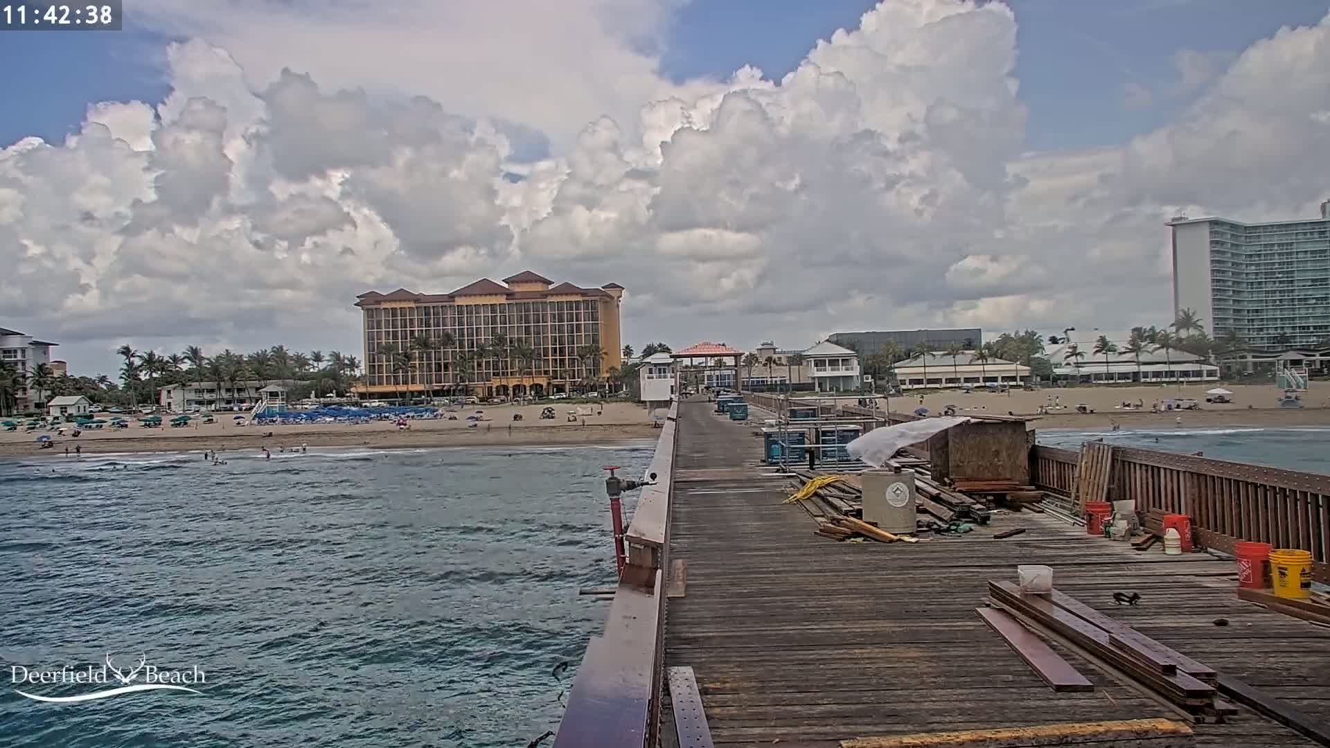 A partially-constructed wooden pier extends over a wavy ocean toward a beach with people and several buildings under a partly cloudy sky.
