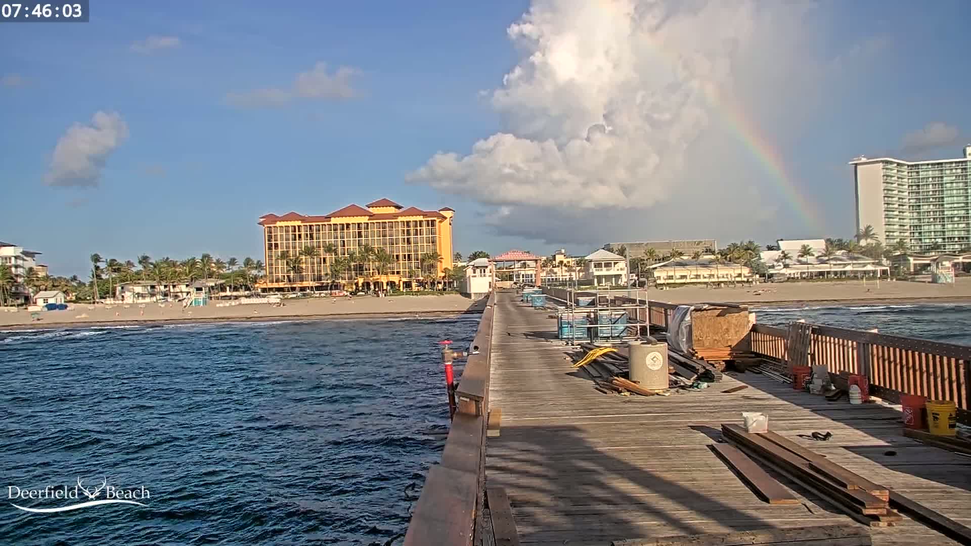 A partly cloudy day with a rainbow shows a view from a pier under construction overlooking a beach with hotels and buildings.
