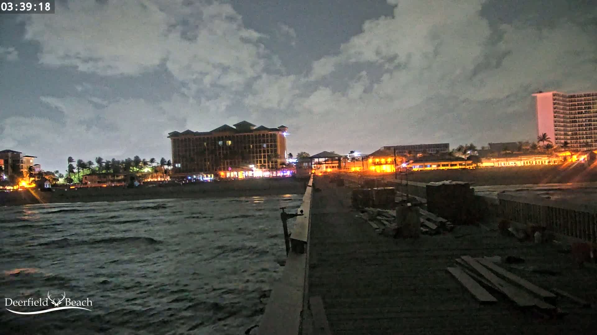 A nighttime view from a pier shows a beach with illuminated buildings and a slightly choppy ocean under a cloudy sky.