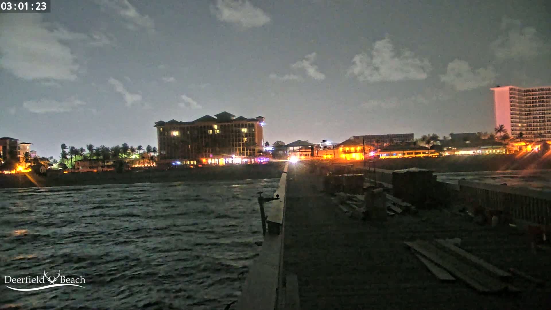 A nighttime view from a pier shows a dark, choppy ocean, illuminated buildings along a shoreline, and partly cloudy skies.