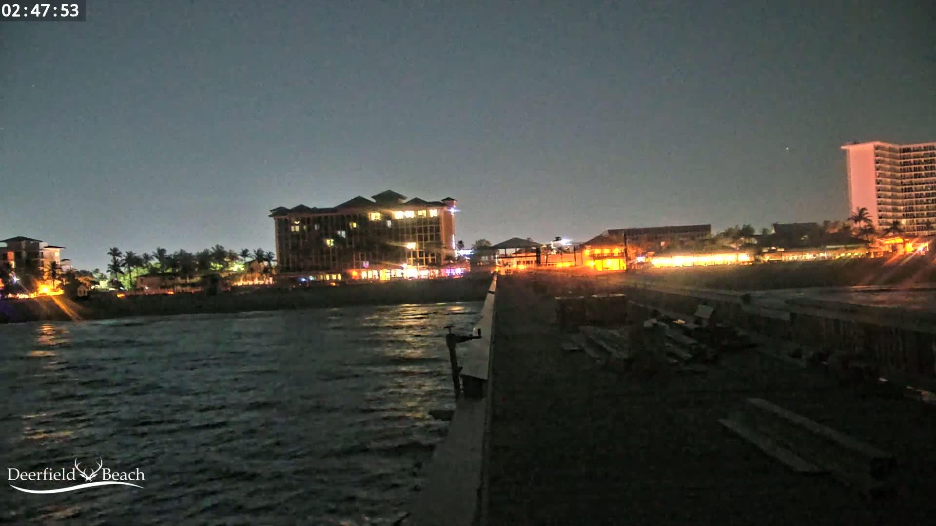 A nighttime view from a pier shows a dark ocean, a beach with illuminated buildings, and a mostly clear sky.