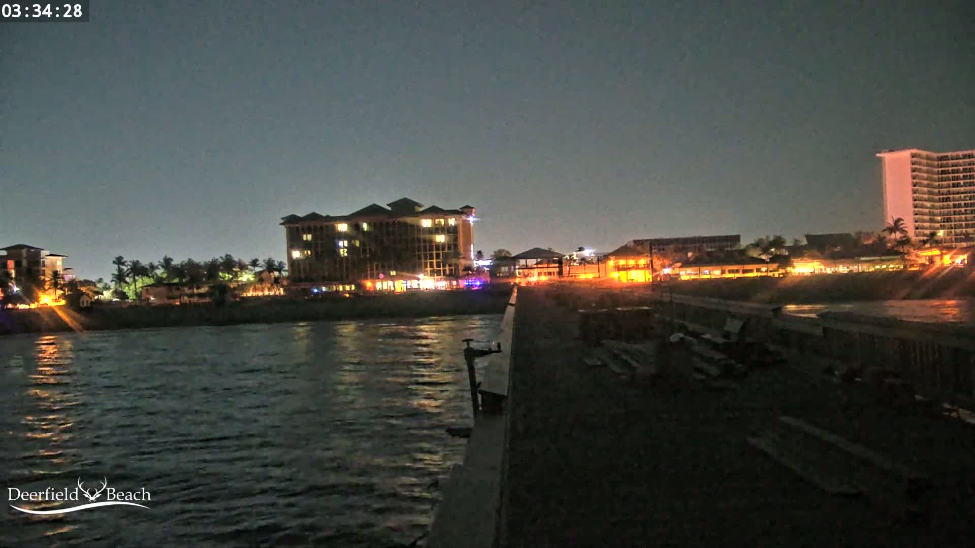 A nighttime view from a pier shows a calm body of water reflecting illuminated buildings along the shore.