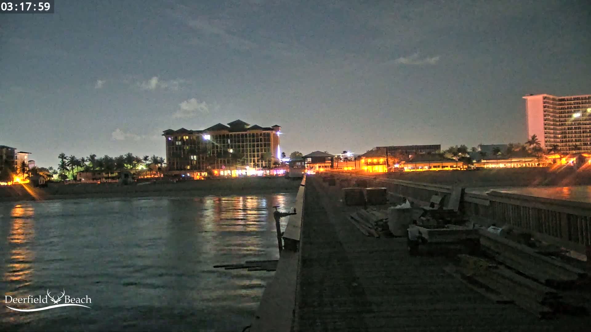 A nighttime view from a pier shows a calm body of water reflecting the lights of buildings along a beach under a mostly clear, dark sky.