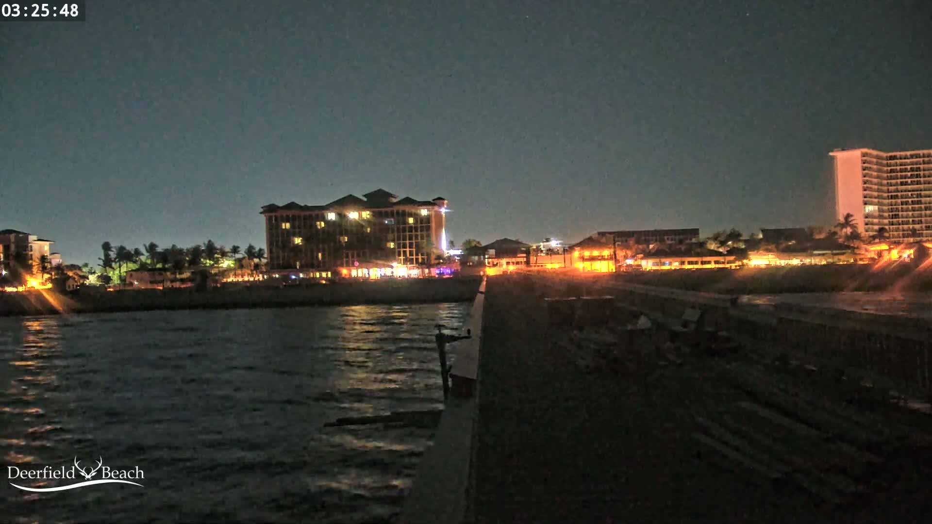 A nighttime view from a pier shows a calm, dark body of water reflecting the lights of nearby buildings, including a large hotel and a high-rise.