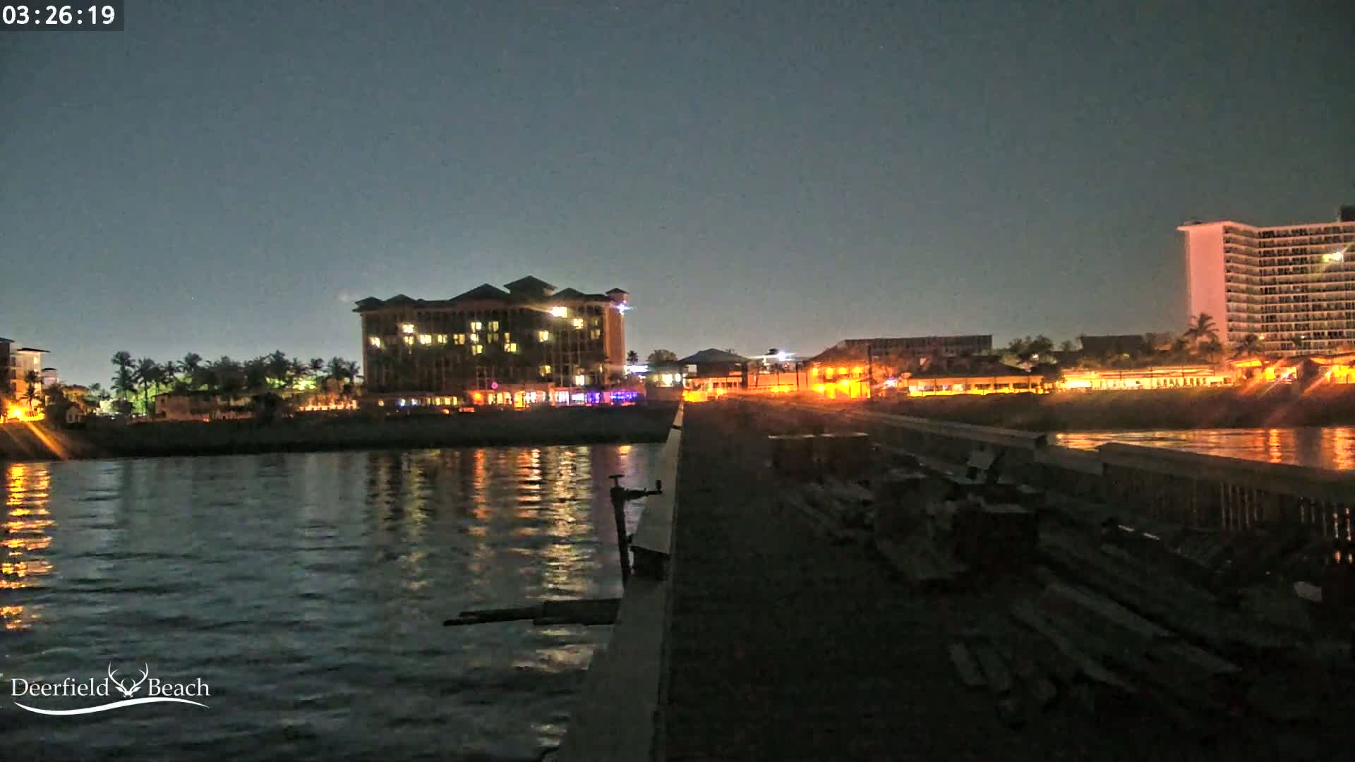 A nighttime view from a pier shows a calm body of water reflecting illuminated buildings along the shore under a dark sky.