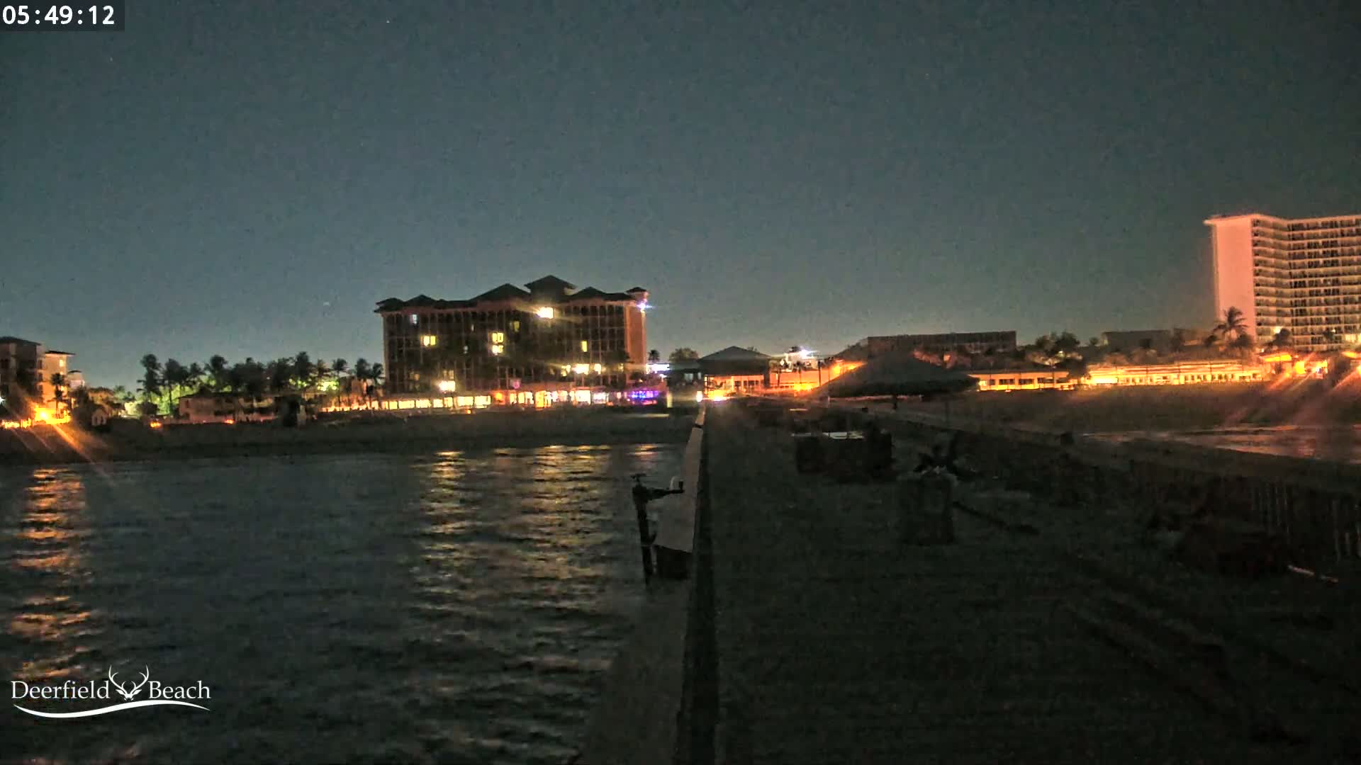 A nighttime view from a pier shows a calm body of water reflecting illuminated buildings along the shore under a dark sky.