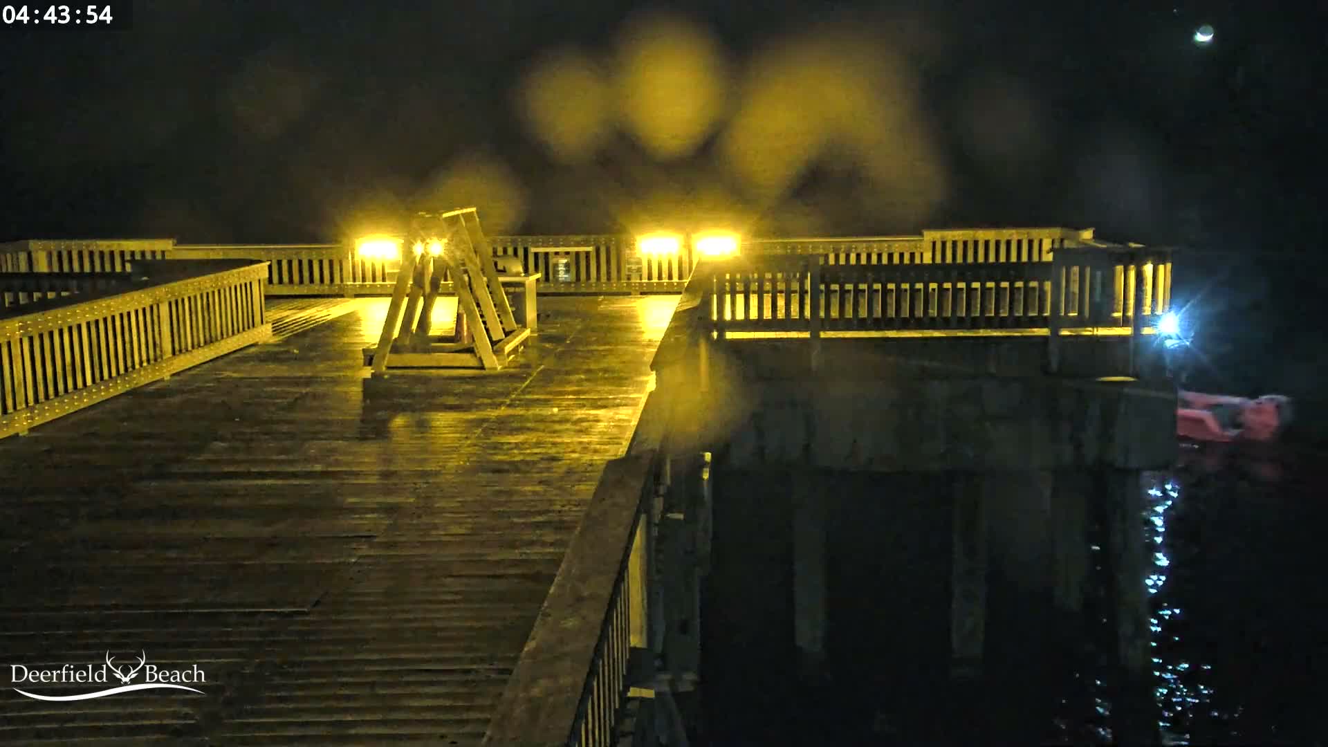 A wet, brightly lit wooden pier extends over dark, misty waters at night, with a person in a red flotation device visible in the water and a faint crescent moon in the sky.