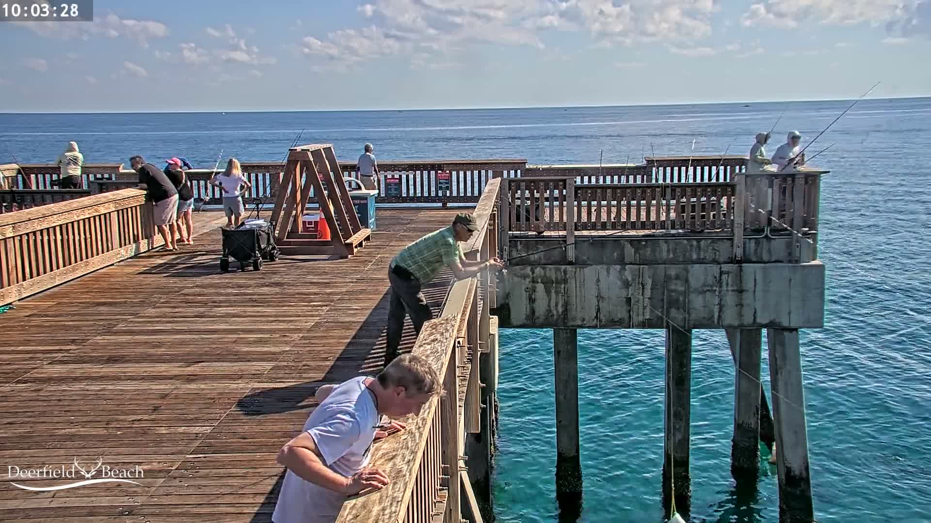 A wet, brightly lit wooden pier extends over dark, misty waters at night, with a person in a red flotation device visible in the water and a faint crescent moon in the sky.