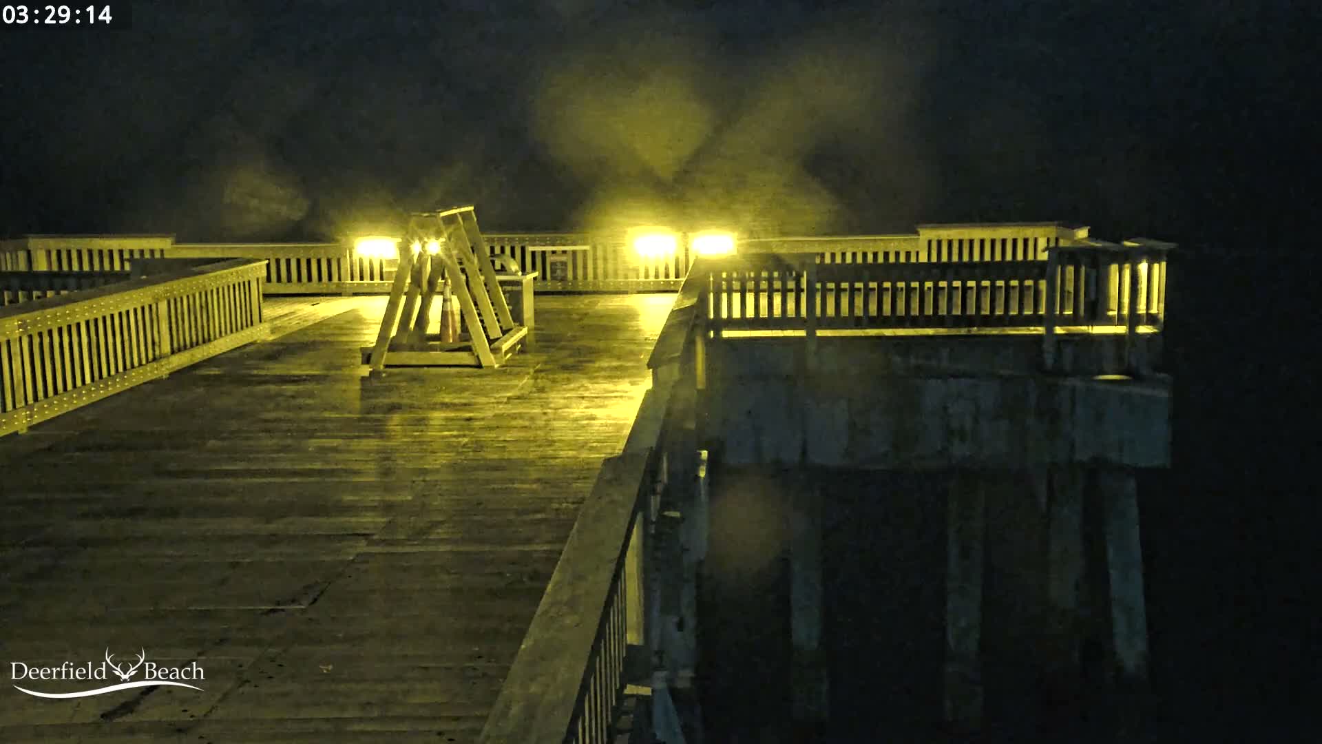 A wet wooden pier is brightly illuminated by yellow lights at night, with a misty atmosphere visible around the light sources.