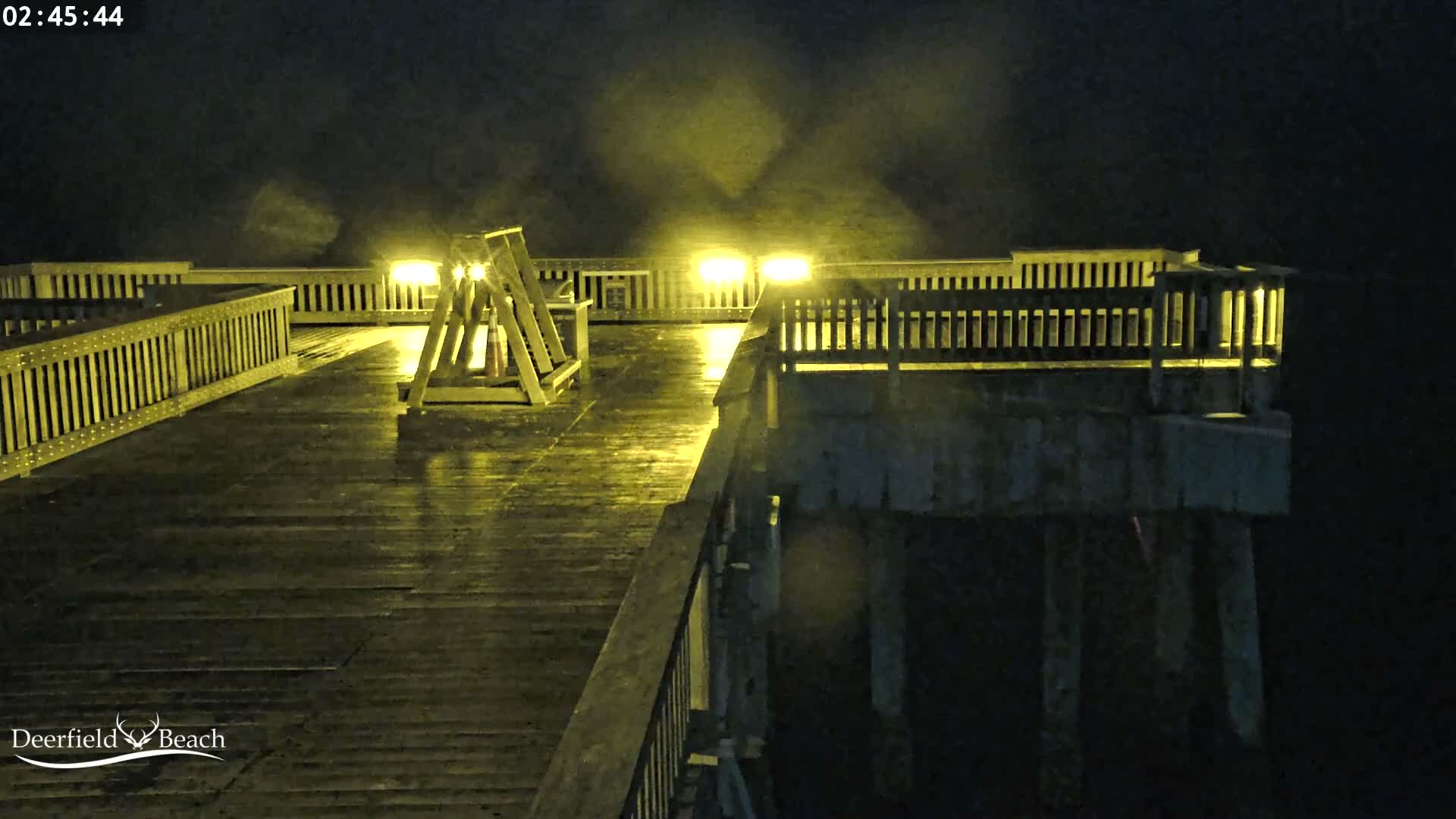 A wet wooden pier with railings and bright overhead lights stretches into the dark night, with light mist visible in the air over the water.