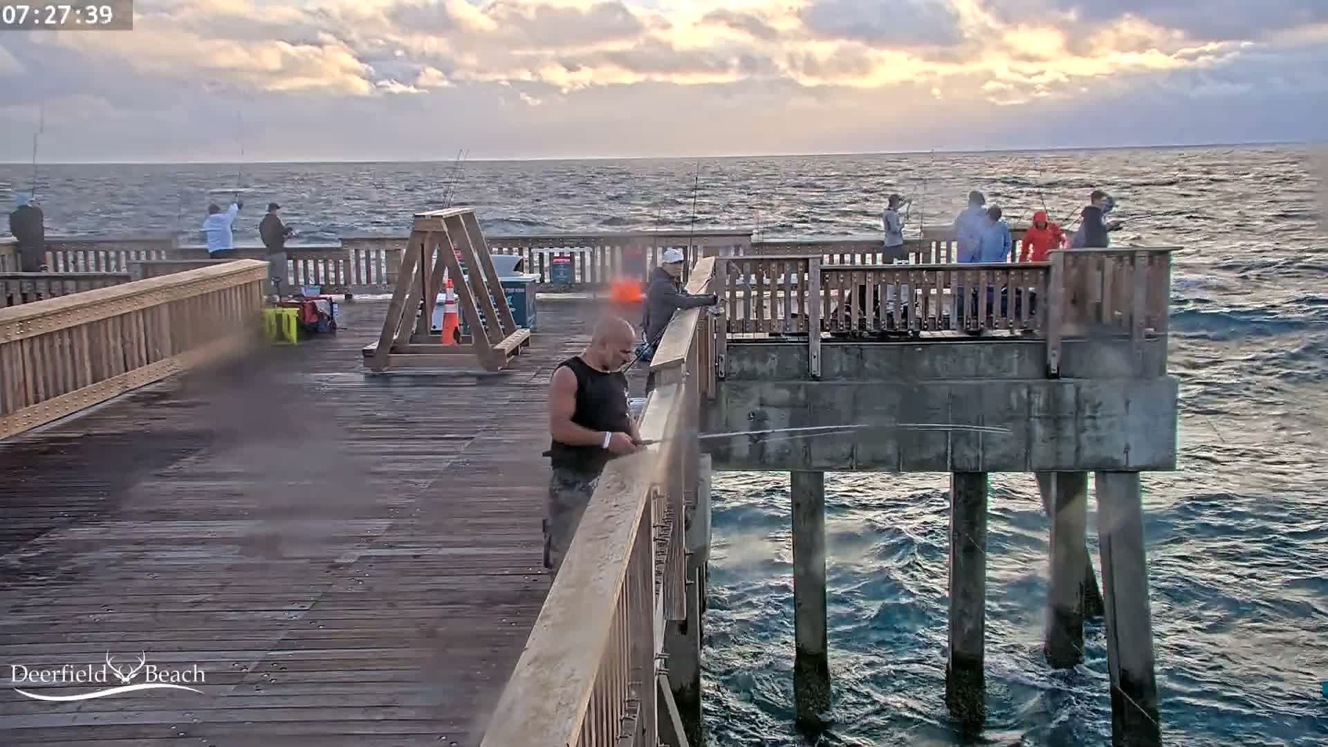 A wet wooden pier with railings and bright overhead lights stretches into the dark night, with light mist visible in the air over the water.
