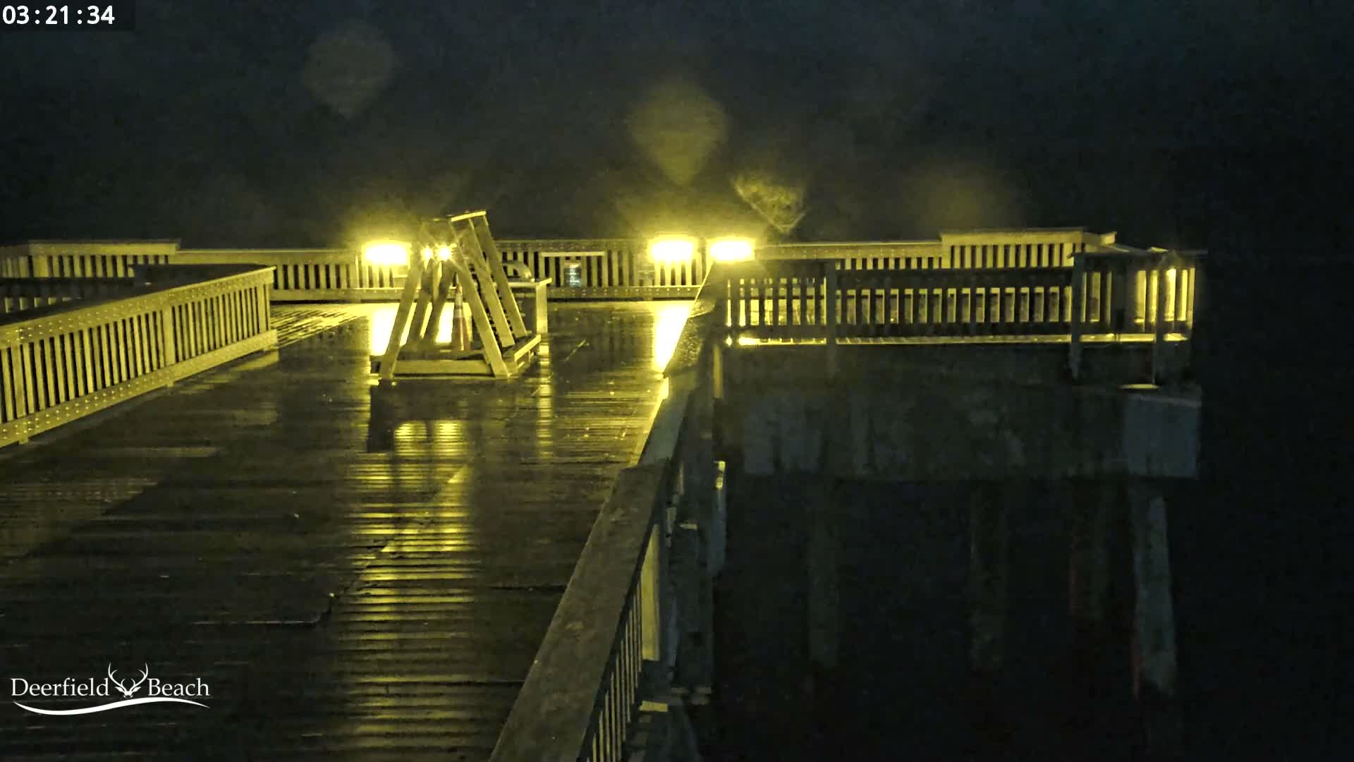 A wet wooden pier, illuminated by bright yellow lights reflecting off its surface, extends over dark water on a rainy night.