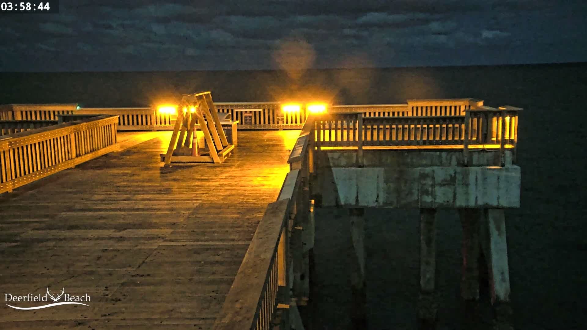 A brightly lit wooden pier, glistening from what appears to be recent rain, extends over dark water under a heavily overcast night sky.