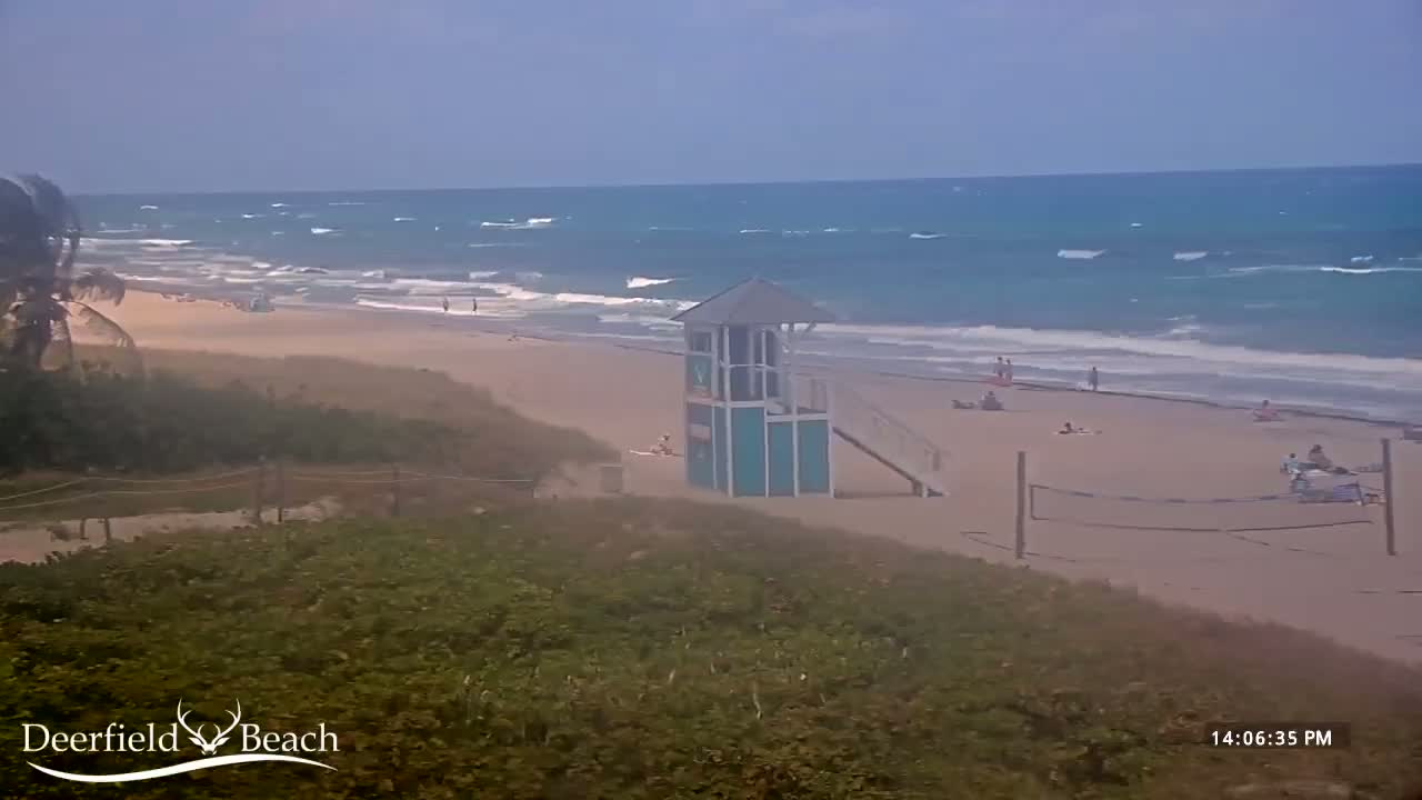 A beach scene shows a lifeguard stand on a sandy beach with a volleyball net and a few people, with somewhat choppy ocean waves visible in the background under a hazy sky.