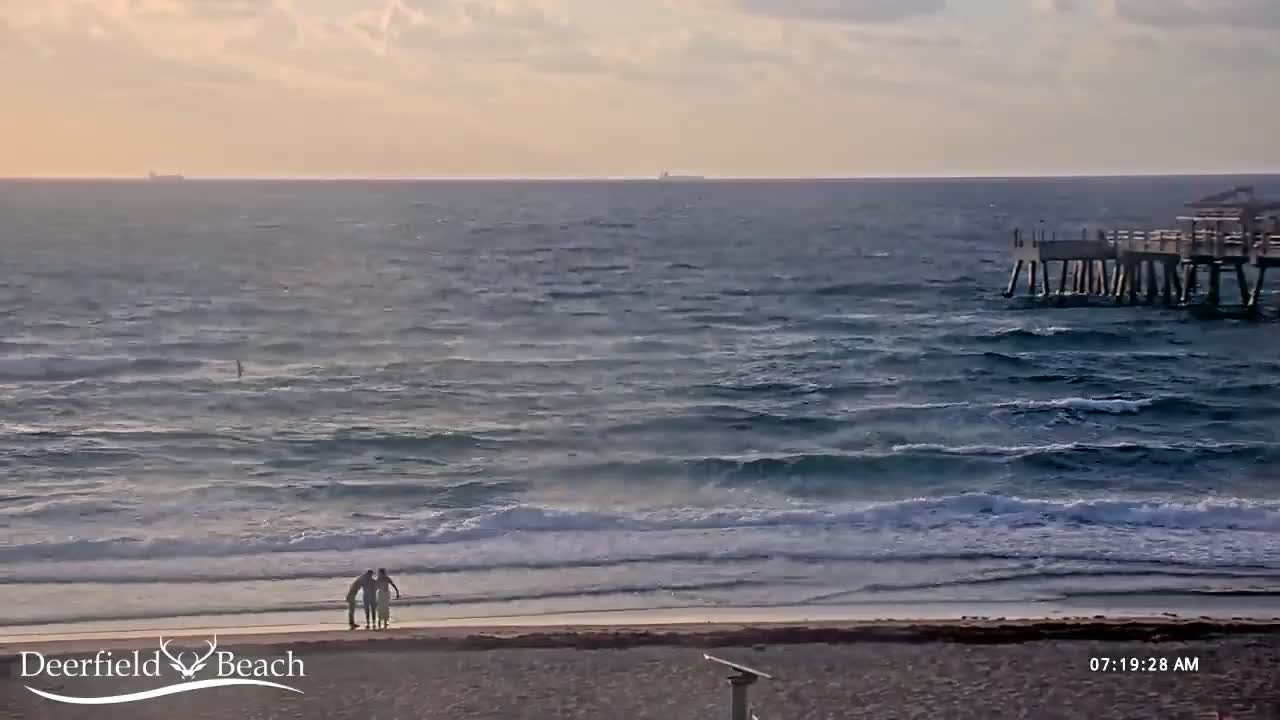 A mostly cloudy sunrise illuminates a beach scene with moderate waves crashing on the shore, a pier in the distance, and three people standing near the water's edge.
