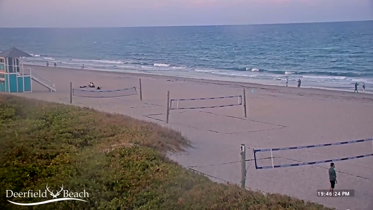 A sandy beach with volleyball nets, a lifeguard stand, and a few people is seen under a partly cloudy sky with a calm ocean in the background.
