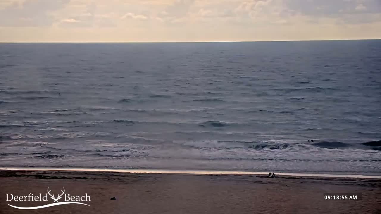 A mostly calm ocean with small waves rolls onto a sandy beach where two people are sitting.