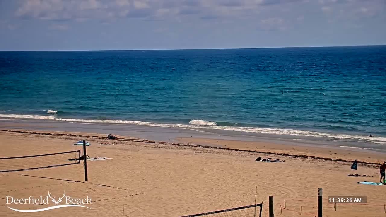 A mostly sunny day at a beach shows a calm ocean with small waves lapping a sandy shore where a few people are relaxing near volleyball nets.