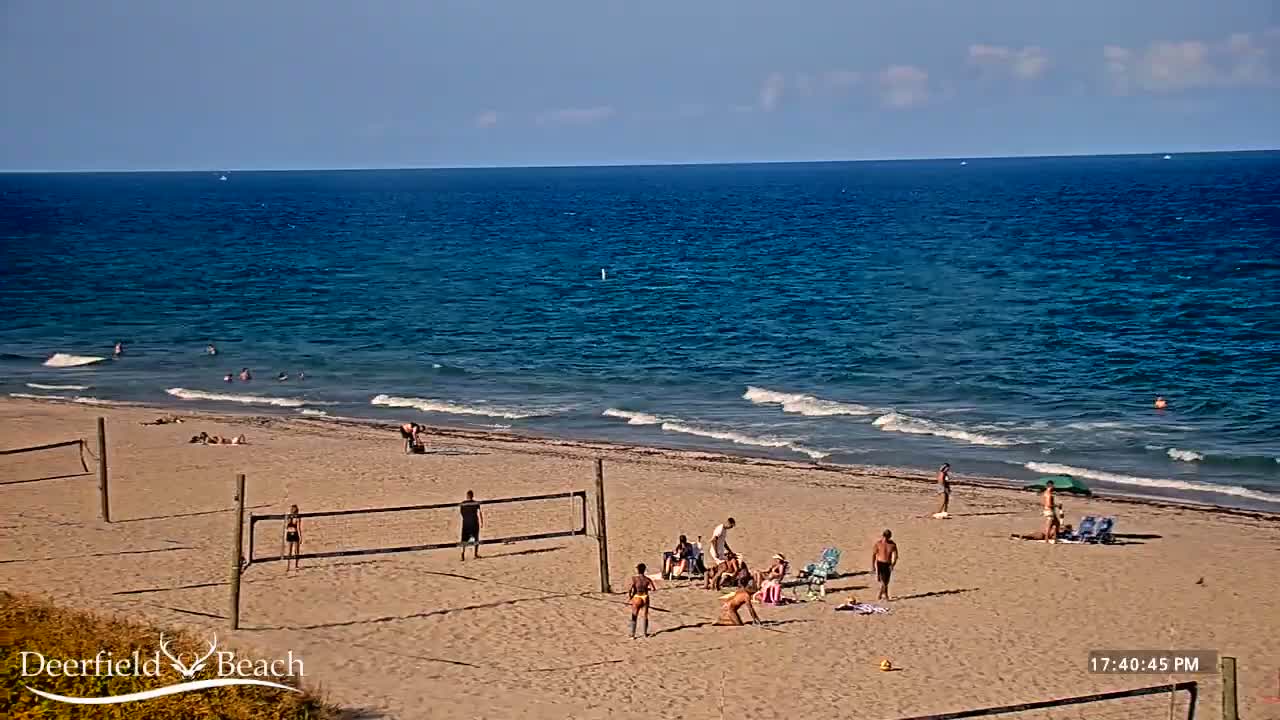 A sandy beach with volleyball nets and several people swimming, sunbathing, and playing volleyball, under a mostly sunny sky with a calm ocean in the background.