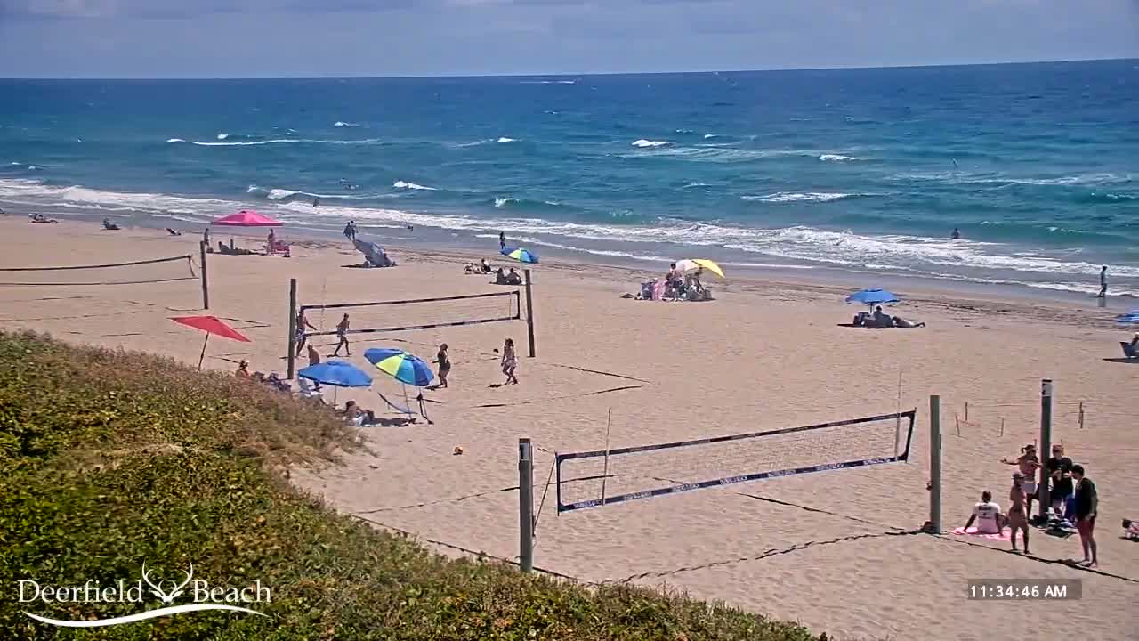A sandy beach with several volleyball nets, people playing volleyball and relaxing under umbrellas, and a wavy ocean under a mostly sunny sky.