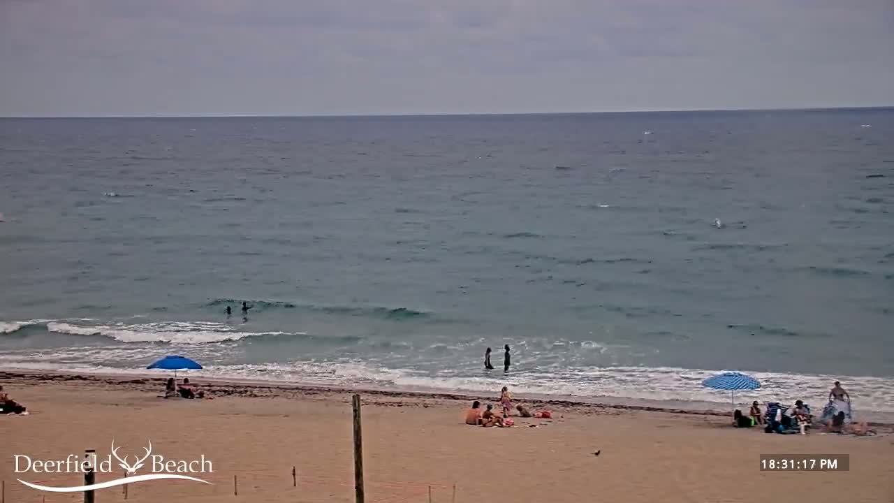 A sandy beach with a few people and umbrellas under an overcast sky, and a calm ocean with small waves.