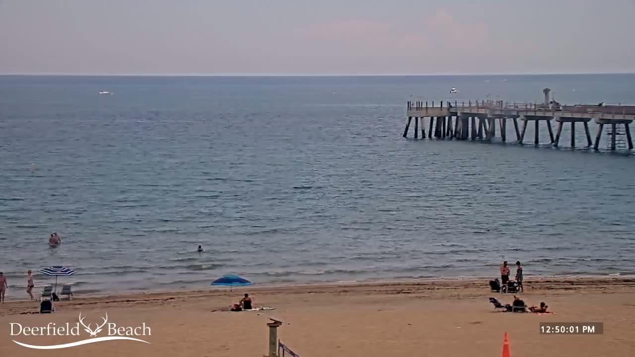 A sandy beach with several people relaxing and swimming in calm ocean water, under a partly cloudy sky, with a pier extending into the ocean in the background.