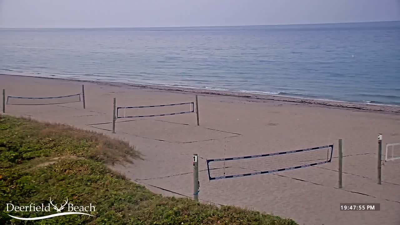 Three volleyball nets are set up on a sandy beach next to a calm ocean under an overcast sky.