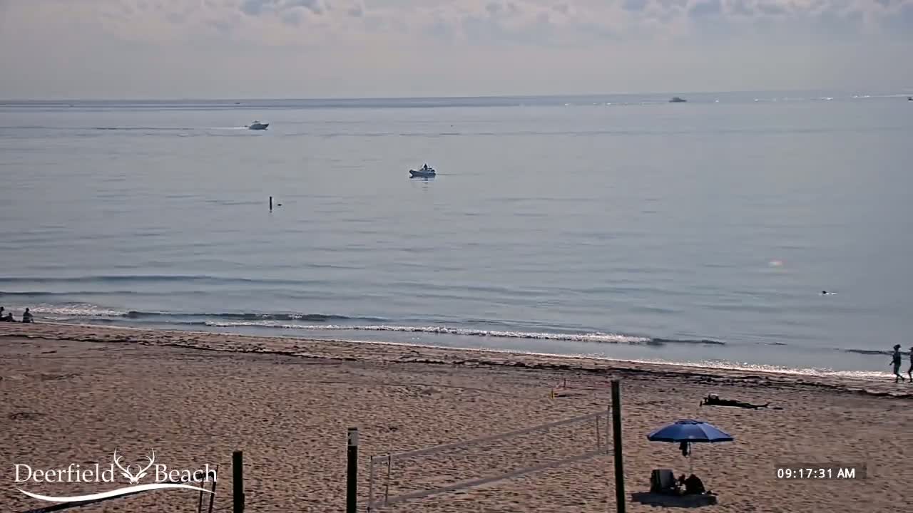 A sandy beach with a volleyball net, a few people, and a blue umbrella is shown with calm ocean waves under a mostly cloudy sky; several boats are visible on the water in the distance.