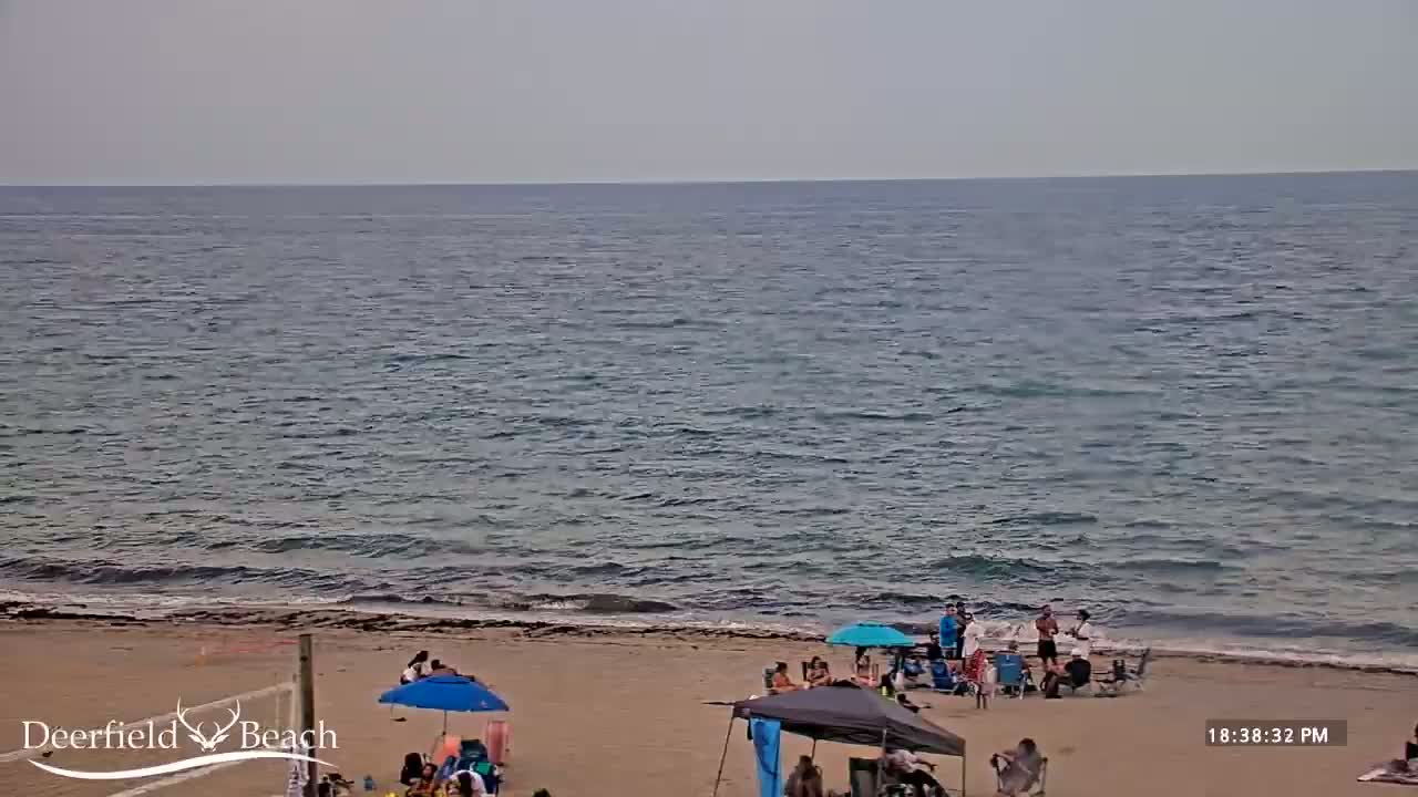 A sandy beach with a few people and beach umbrellas under a partly cloudy sky, overlooks a calm, dark-blue ocean with small waves.