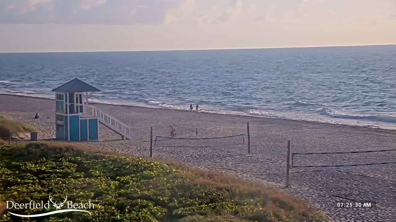 A mostly calm ocean with small waves laps a sandy beach where a lifeguard stand and volleyball net are visible, under a partly cloudy sky.