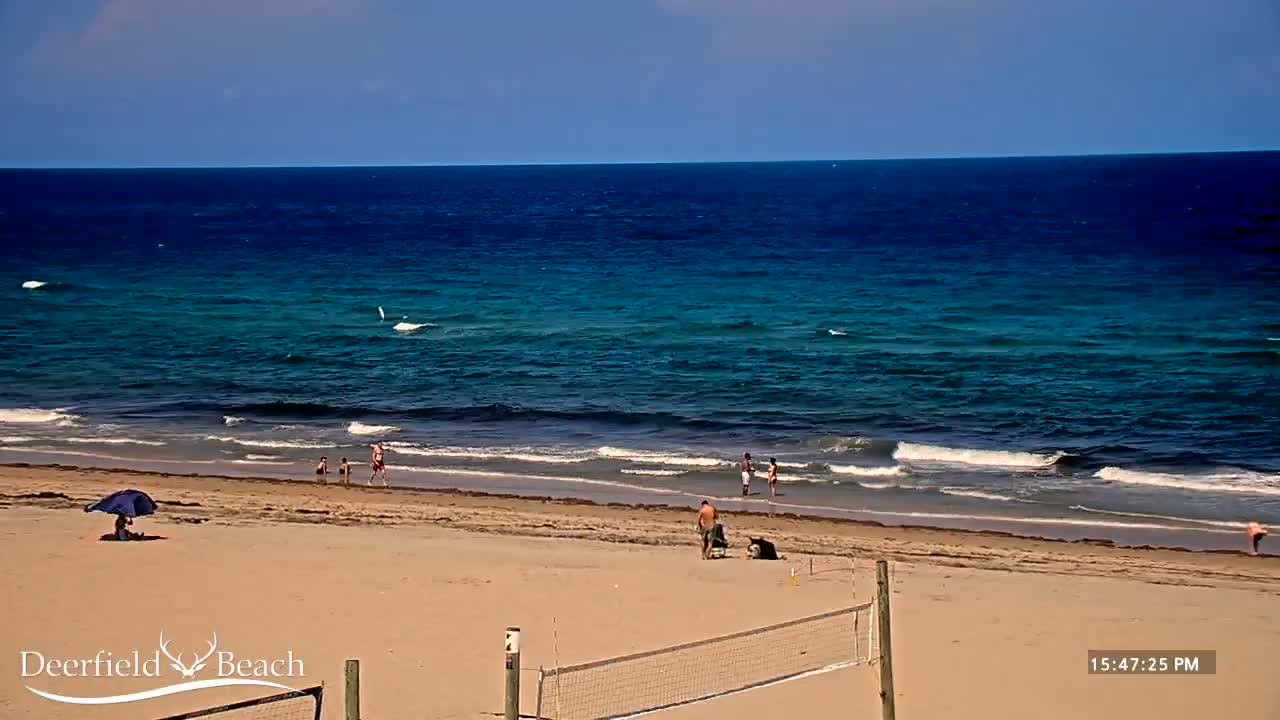 A sandy beach with a few people and an empty volleyball net is visible, under a clear sky, next to a calm, blue ocean with small waves.