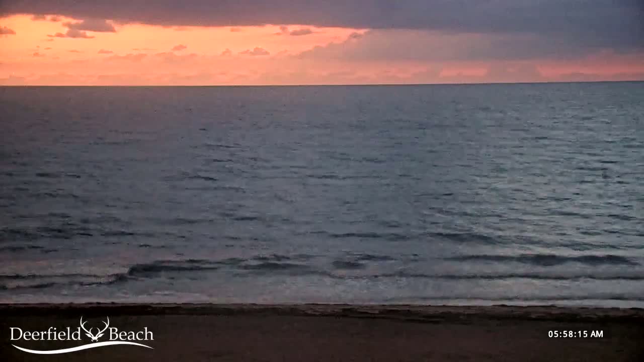 A dark ocean with small waves meets a sandy beach under a sunrise sky with pink and purple clouds.