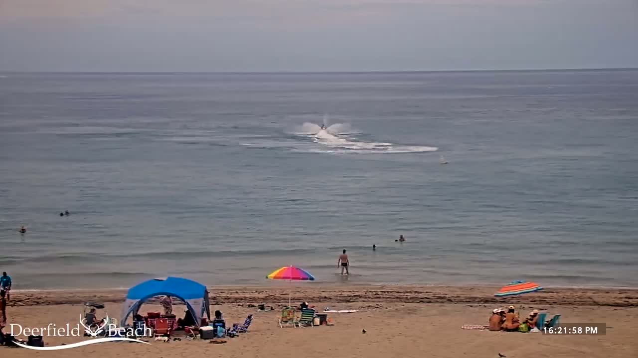 A sandy beach with several people and beach umbrellas, with a jet ski creating a spray in the ocean under an overcast sky.